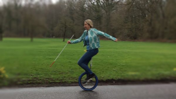 Auf dem Bild sieht man Dörte Maack auf einem Einrad fahren. Mit dem linken Arm hält sie das Gleichgewicht, in der rechten Hand hält sie ihren Blindenstock. Im Hintergrund ist eine große Grünfläche mit Bäumen zu sehen.